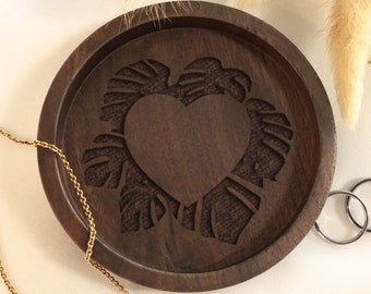 May include: A round, dark brown wooden tray with a carved heart and leaf design. A gold necklace and two silver rings are near the tray. Dried plants are in the background. The tray is a decorative item.