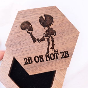 May include: A wooden hexagonal box with a black velvet interior. The lid features an engraved image of a character holding a skull and the text "2B or not 2B".