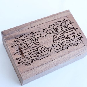 Puede incluir: Una caja de madera con un corazón y un diseño de circuito impreso grabado en la parte superior. La caja está hecha de madera oscura y tiene un acabado natural.