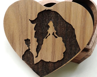 May include: A wooden heart-shaped box with a hinged lid. The box is decorated with a silhouette of Belle and the Beast from the Disney movie Beauty and the Beast. The silhouette is carved into the wood and is painted black.