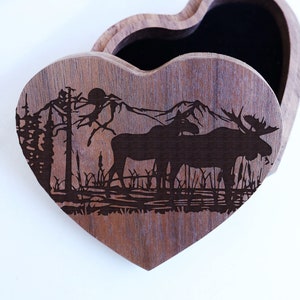 May include: A wooden heart-shaped box with a hinged lid. The box is decorated with a laser-engraved design of two moose standing in a forest setting. The box is open, revealing a dark interior.