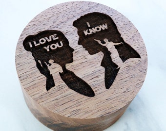 May include: A round, wooden box with a laser-etched design. The design features two silhouettes facing each other, with the words "I LOVE YOU" and "I KNOW" etched into the wood. The box has a natural wood grain finish.