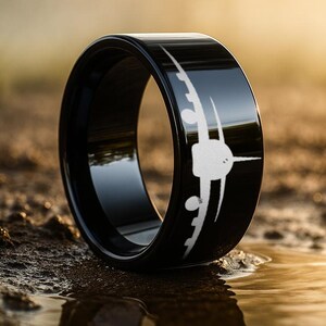 May include: A black ring with a polished finish, featuring a white airplane design. The ring is set against a blurred outdoor background, highlighting the ring's reflective surface and the detailed aircraft graphic. The ring is a jewelry item.