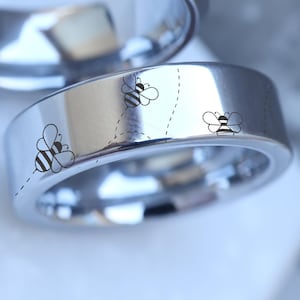 May include: A shiny, silver ring with a flat band. The ring is decorated with three small, black and white bee illustrations, each with a dotted flight path. The ring is likely made of stainless steel or a similar metal.