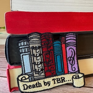 Könnte beinhalten: Gestickter Aufnäher mit einem Stapel bunter Bücher und einem Banner mit der Aufschrift "Death by TBR..." Der Aufnäher befindet sich auf einer Holzoberfläche.