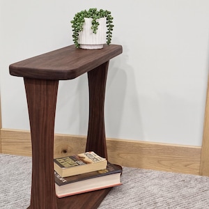 May include: A dark brown wooden side table with a rectangular top and curved legs. A white ceramic pot with a green plant sits on top. Two books are stacked on the lower shelf. The table is approximately 30 inches tall.