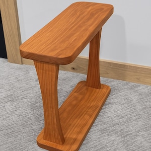 May include: A handcrafted, cherry-wood side table with a curved top and base, supported by two elegantly curved legs. The table has a warm, reddish-brown finish, showcasing the natural wood grain. The design is modern and minimalist.
