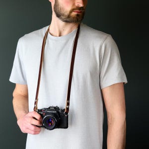 May include: A dark brown leather camera strap attached to a black Pentax camera with a lens. The strap features silver-toned metal rivets securing the leather loops to the camera body, providing a functional way to carry the photographic equipment.
