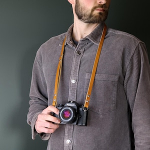 May include: A tan leather camera strap with silver-toned metal rivets and circular rings attached to a black Nikon camera. The strap features a smooth, flat design that hangs around the neck, securing the camera body with a lens attached.
