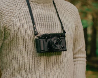 May include: A black camera with a leather strap hanging around a person's neck. The camera has a silver lens and a black body.