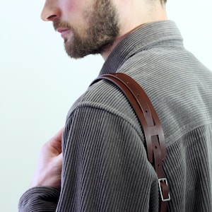 Può includere: Un cinturino in pelle marrone con dettagli in argento è indossato sulla spalla di una persona che indossa una camicia di velluto a coste grigia.