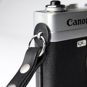 May include: A black leather camera strap attached to a silver and black Canon QL camera body. The strap features a circular black leather protective pad, a silver metal split ring, and two silver metal rivets securing the leather loop together.