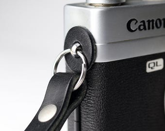 May include: A black leather camera strap attached to a silver and black Canon QL camera body. The strap features a circular black leather protective pad, a silver metal split ring, and two silver metal rivets securing the leather loop together.