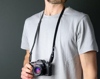 May include: A black Pentax camera with a lens attached, suspended from a thin black leather neck strap. The camera body is rectangular with a prominent lens barrel, and the strap features metal rivets at the connection points to the camera.