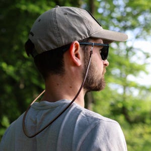 May include: A person wearing a gray baseball cap, sunglasses, and a brown leather eyeglass strap. The strap is attached to the glasses and hangs down. The background is a blurred view of green trees.