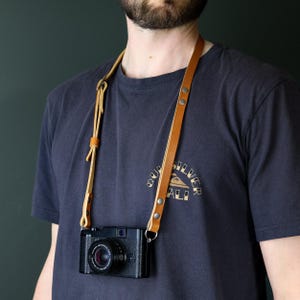 May include: A tan leather camera strap featuring adjustable buckles and silver-toned metal rivets, attached to a black digital camera. The strap is worn around the neck, showcasing the smooth, light brown leather construction and secure metal hardware.
