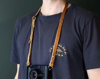 May include: A tan leather camera strap featuring adjustable buckles and silver-toned metal rivets, attached to a black digital camera. The strap is worn around the neck, showcasing the smooth, light brown leather construction and secure metal hardware.