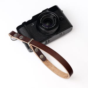 May include: A black digital camera with a textured body and a Fujinon Aspherical Lens, attached to a dark brown leather wrist strap. The strap features silver-toned metal rivets and a rectangular metal buckle, creating a secure loop for the camera.