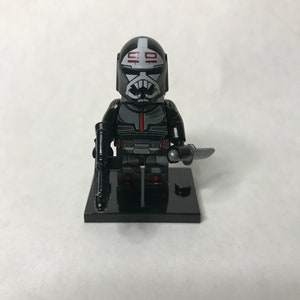 Könnte beinhalten: Eine Lego-Minifigur eines Charakters in einem schwarz-grauen Anzug, der einen Blaster und ein Messer hält. Die Figur hat einen Helm mit weißen Markierungen und einem roten Streifen. Sie steht auf einer schwarzen Basis.