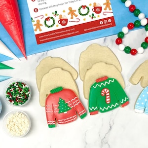 May include: A Christmas cookie decorating kit with four undecorated sugar cookies shaped like sweaters, three containers of sprinkles, four piping bags, and a string of red and green beads. The cookies are ready to be decorated with icing and sprinkles.