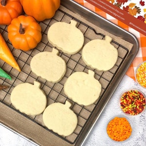 May include: A close-up of a baking sheet with a wire rack holding seven pumpkin-shaped sugar cookies. The cookies are un-decorated and ready to be decorated. There are three bowls of sprinkles and a few pumpkins in the background.