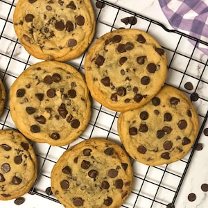 May include: Overhead shot of freshly baked chocolate chip cookies on a black wire cooling rack. The cookies are golden brown with visible chocolate chips. A purple and white checkered cloth is in the background, along with scattered chocolate chips.