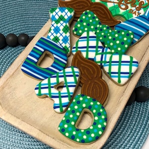 May include: A set of decorated sugar cookies spelling out "DAD" with a bow tie, mustache, and tie. The cookies are decorated with green, blue, and white plaid, polka dots, and stripes. The cookies are on a wooden tray with a "Happy Father's Day" sign.