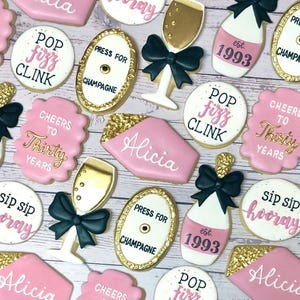 May include: A set of 14 decorated sugar cookies with pink, gold, and black frosting. The cookies feature various designs, including champagne bottles, glasses, and the words "Pop Fizz Clink", "Cheers to Thirty Years", and "Alicia".