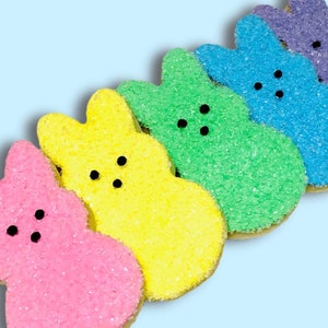 May include: Five pastel-colored bunny-shaped cookies decorated with sugar sprinkles. The cookies are pink, yellow, green, blue, and purple.