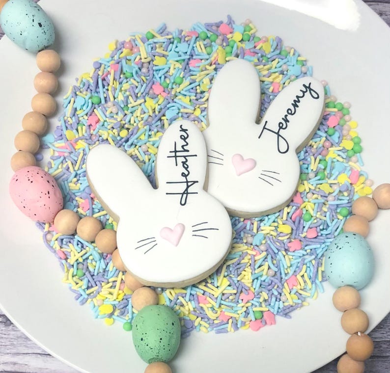 May include: White bunny-shaped cookies with the names "Heather" and "Jeremy" written on them, decorated with pink hearts and whiskers. The cookies are surrounded by colorful sprinkles and Easter-themed beads.