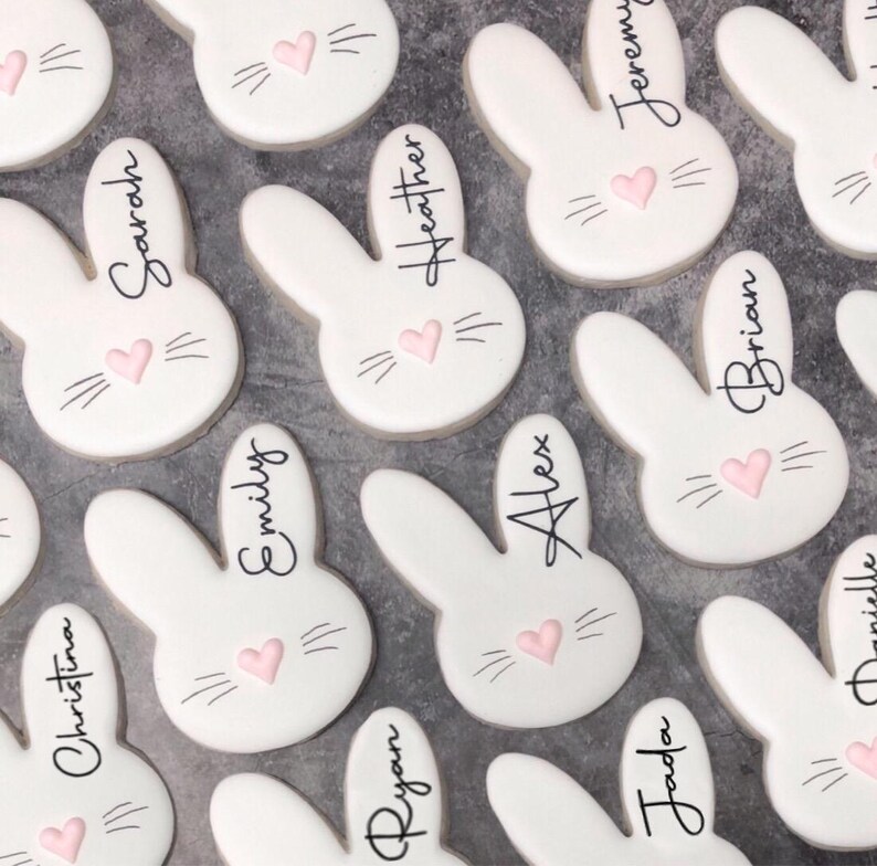 May include: White bunny-shaped cookies with pink heart noses and black whiskers. Each cookie has a name written in black script, including "Sarah", "Heather", "Jeremy", "Brian", "Emily", "Alex", "Christina", "Ryan", and "Jada".