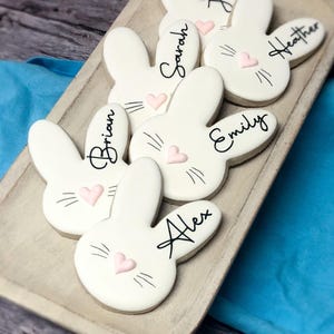 May include: White bunny-shaped cookies with black whiskers, pink heart noses, and names written in black script. The cookies are arranged on a wooden tray, perfect for Easter or a spring celebration.