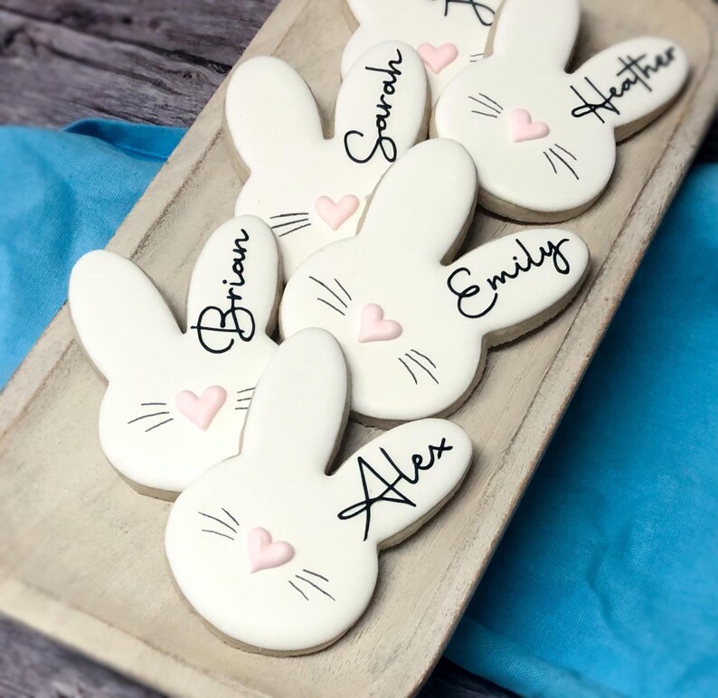 May include: White bunny-shaped cookies with black whiskers, pink heart noses, and names written in black script. The cookies are arranged on a wooden tray, perfect for Easter or a spring celebration.