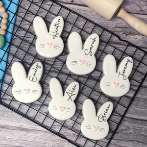 May include: Six white bunny-shaped cookies with black whiskers, names, and pink heart accents are arranged on a black wire rack. A wooden rolling pin rests nearby. A beaded garland with an Easter egg is in the upper left corner.