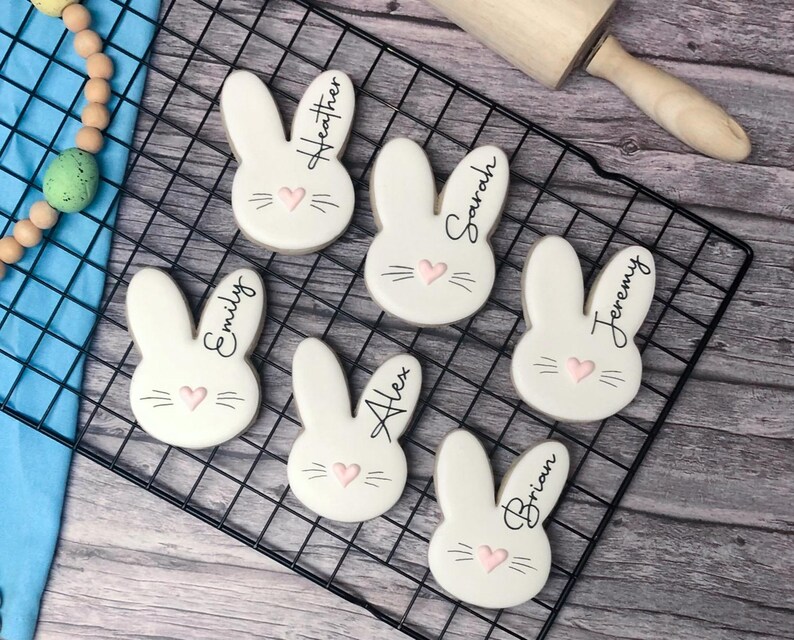 May include: Six white bunny-shaped cookies with black whiskers, names, and pink heart accents are arranged on a black wire rack. A wooden rolling pin rests nearby. A beaded garland with an Easter egg is in the upper left corner.