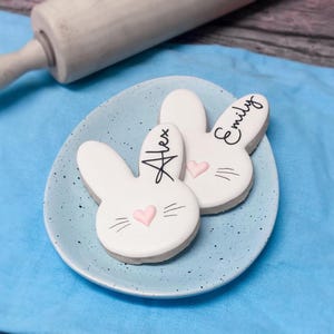 May include: Two bunny-shaped cookies with white icing, pink heart noses, and black whiskers sit on a light blue plate. One cookie has the name "Alex" and the other "Emily" written in black. A wooden rolling pin is in the background.