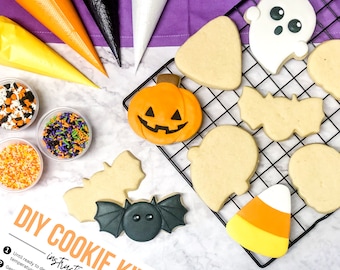 May include: DIY cookie kit with a variety of Halloween-themed cookies, including a ghost, a bat, a pumpkin, a candy corn, and a triangle. The cookies are on a black wire rack and there are sprinkles and icing bags in the background.