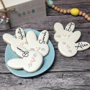 May include: A light blue plate holds bunny-shaped cookies with names written on them. The cookies are white with pink heart noses and black whiskers. The names are written in black script. The background includes a wooden surface and a decorative wooden sign that says "Bunny Kisses and Easter Wishes."