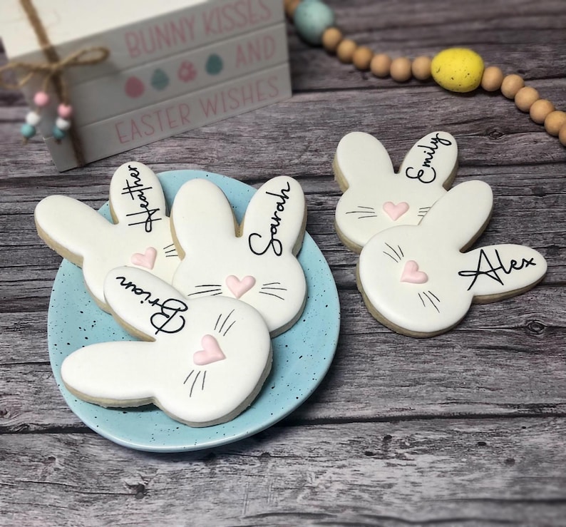 May include: A light blue plate holds bunny-shaped cookies with names written on them. The cookies are white with pink heart noses and black whiskers. The names are written in black script. The background includes a wooden surface and a decorative wooden sign that says "Bunny Kisses and Easter Wishes."