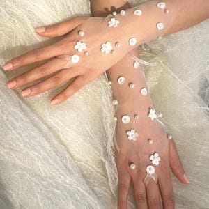 Könnte beinhalten: Transparente weiße Handschuhe, verziert mit kleinen weißen Blumen und Perlen. Die Handschuhe haben eine Fingerschlaufe und eignen sich für einen besonderen Anlass, wie eine Hochzeit oder eine formelle Veranstaltung.
