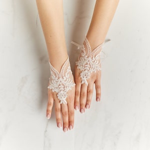 May include: Ivory lace fingerless gloves with detailed floral and scrollwork patterns. The gloves have a delicate, open design and are tied with a small ribbon. These elegant gloves are suitable for weddings or special events.