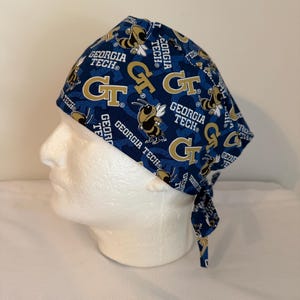 May include: A navy blue scrub cap featuring a repeating pattern of gold "GT" logos, yellow jackets, and the words "Georgia Tech". The cap is tied at the back.