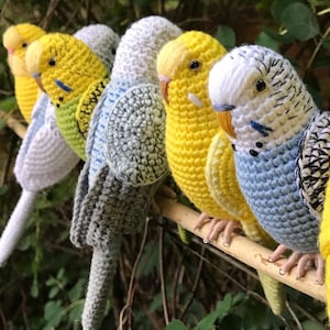 May include: A row of six colorful, crocheted parakeets perched on a wooden branch. The birds feature various colors, including yellow, blue, white, and green. The parakeets are handmade and have detailed features.