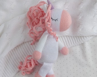 Puede incluir: Un unicornio de peluche blanco tejido a crochet con hilo rosa para la crin y la cola. El unicornio está acostado sobre una manta blanca con un pequeño patrón de puntos blancos.
