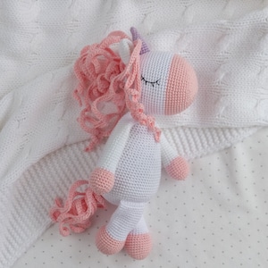 May include: A white crocheted unicorn stuffed animal with pink yarn for the mane and tail. The unicorn is lying on a white blanket with a small white dot pattern.