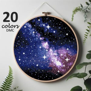 Könnte beinhalten: Ein runder Stickrahmen mit einem Kreuzstich-Design einer Galaxie in Blau-, Lila- und Weißtönen. Der Text "20 Farben DMC" ist in Schwarz dargestellt. Der Rahmen ist aus Holz und hängt an einer weißen Wand.