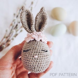 Könnte beinhalten: Ein gehäkeltes Hasenspielzeug mit langen Ohren, einem rosa Blumenband und gestickten Augen. Der Hase ist braun und hat eine rosa Nase.