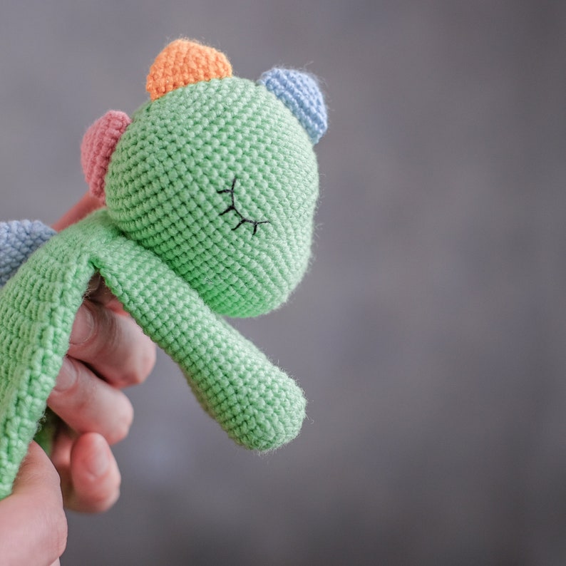 Op de afbeelding: Een groen gehaakt dinosaurus speelgoed met kleurrijke accenten en geborduurde ogen. Het speelgoed wordt vastgehouden door een hand.