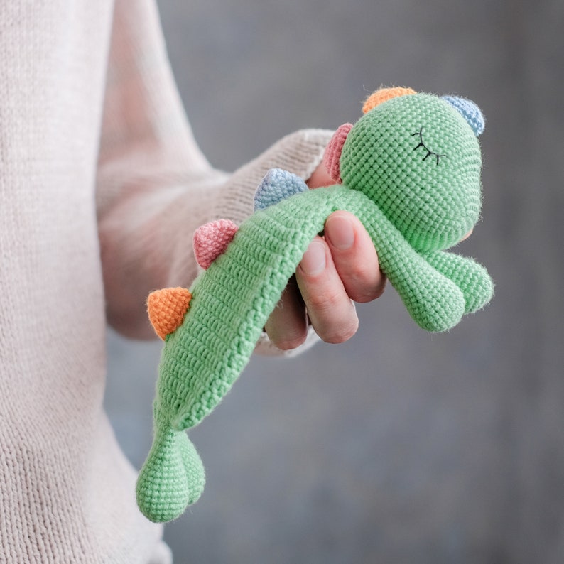 Op de afbeelding: Een groen gehaakt dinosaurus speelgoed met kleurrijke accenten. De dinosaurus heeft gesloten ogen en een slaperige uitdrukking.