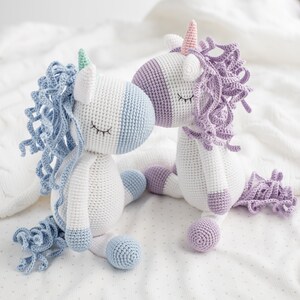 May include: Two crocheted unicorn toys, one with blue and white yarn and the other with purple and white yarn. Both have closed eyes and are sitting on a white blanket.
